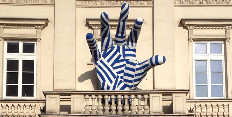 W centralnym punkcie zdjęcia rzeźba dłoni pomalowana w biało-niebieskie pasy. W tle trzy okna. Całość w promieniach słońca