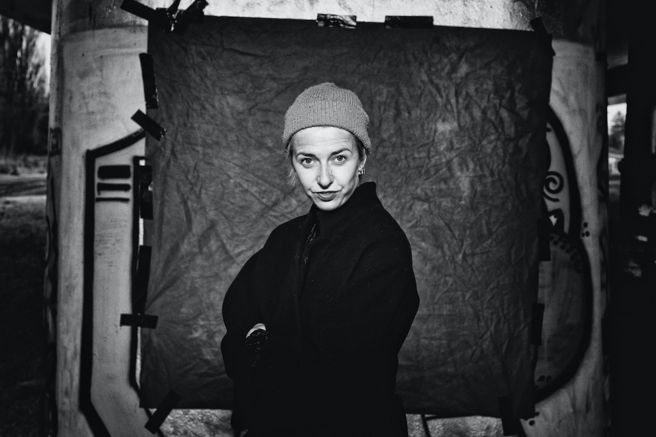 Czarno-biała fotografia, na której znajduje się Justyna Sobczyk. Widzimy ją od pasa w gorę. Ma założone o siebie ręce. Ubrana na czarno. Jest w czapce. Patrzy na nas zadziornie. Stoi na tle szarego prostokąta.