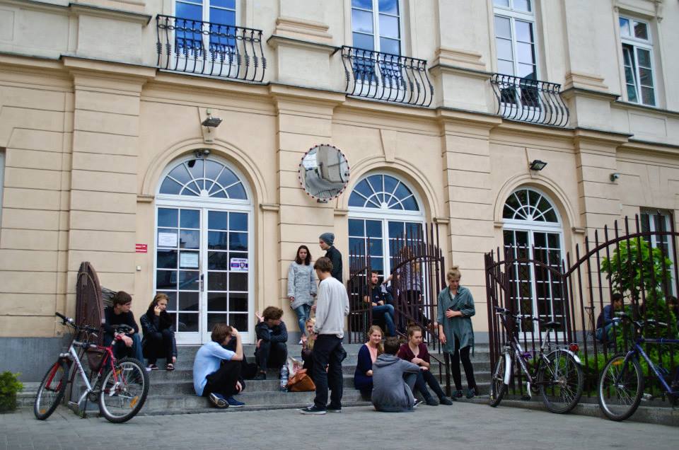 Wejście do budynku Akademii Teatralnej od strony parkingu. Przed wejściem, na schodach, siedzą i stoją młodzi ludzie - czekają na swój egzamin. Wokół nich stoją rowery.