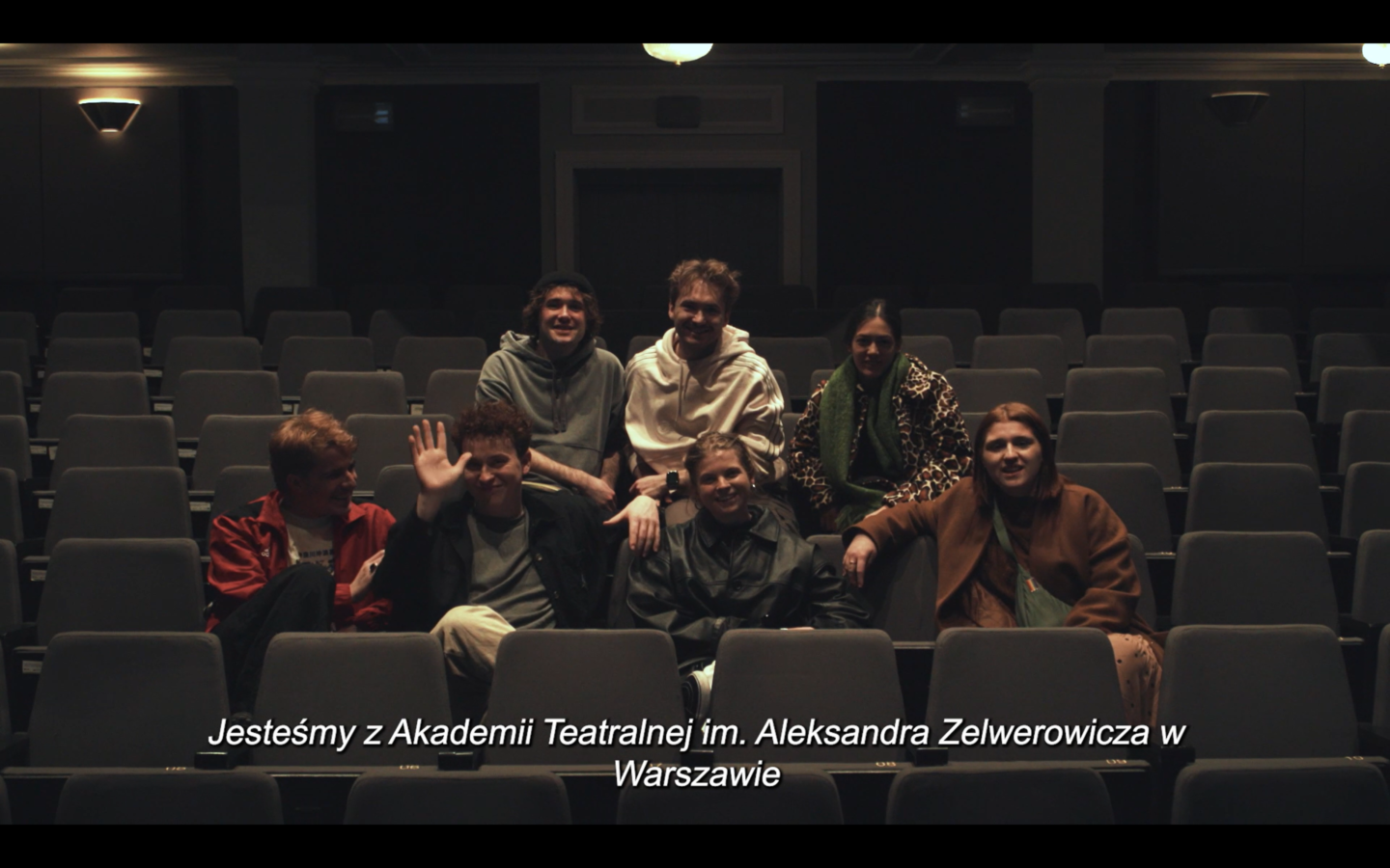 Kadr z filmu. W teatralnej widowni siedzi siedmiu młodych ludzi. Poniżej nich widnieje napis: "Jesteśmy z Akademii Teatralnej im. Aleksandra Zelwerowicza w Warszawie".