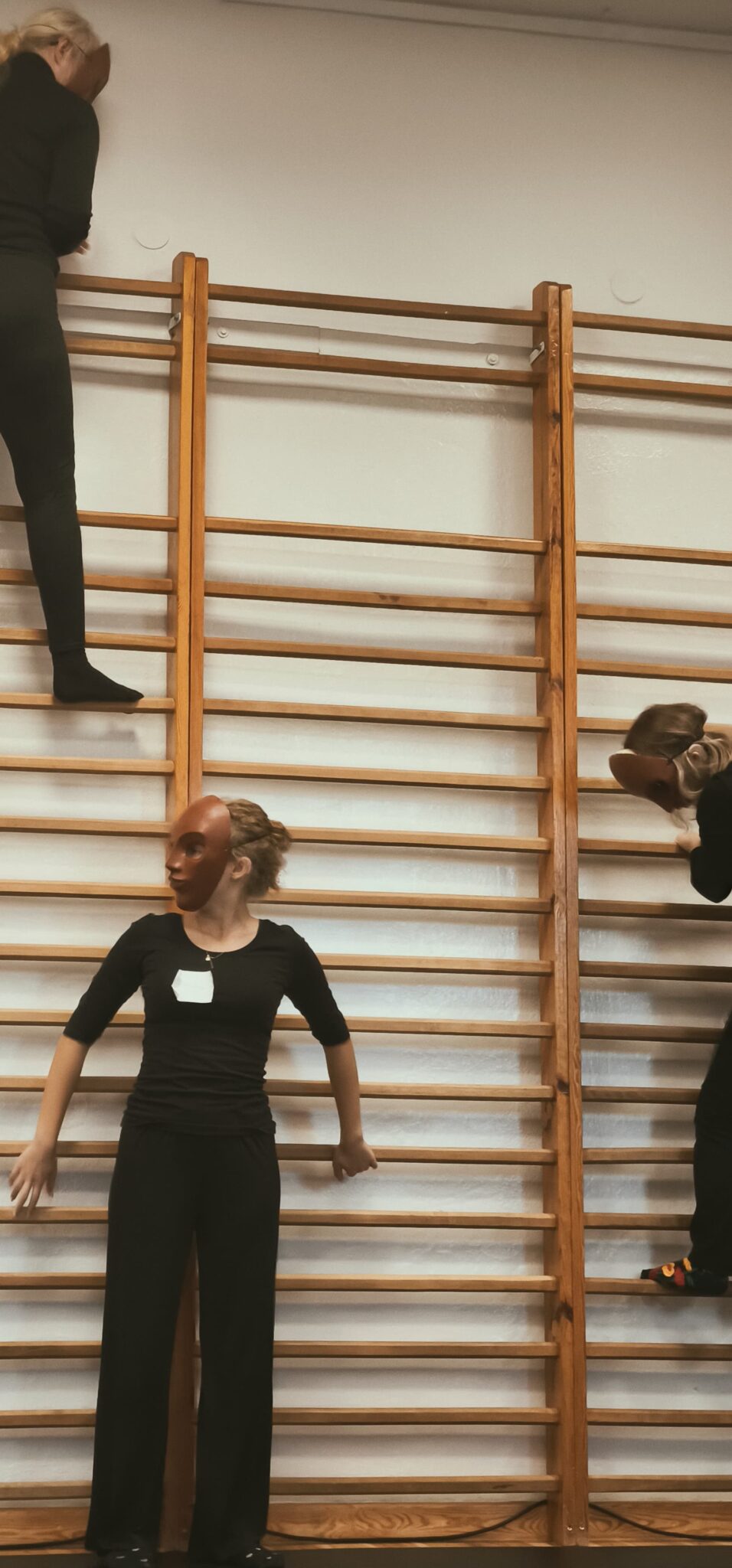 Osoby w maskach teatralnych wspinają się i stoją na drewnianych drabinkach w sali gimnastycznej. Jedna z uczestniczek stoi na środku, oparta o szczebel, spoglądając w bok.
