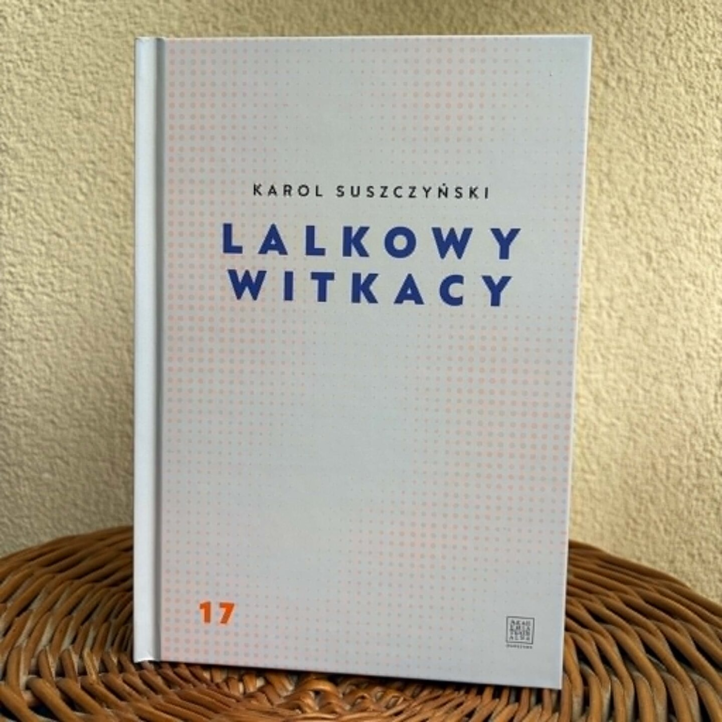 Zdjęcie zrobione na wprost, przedstawiające książkę „Lalkowy Witkacy” (autor Karol Suszczyński) stojącą pionowo. Książka opiera się o chropowatą ścianę o tynku strukturalnym w kolorze beżowym, a jej podstawa znajduje się na okrągłym, brązowym przedmiocie z wiklinowej plecionki. Wyraźnie widać całą przednią okładkę publikacji.