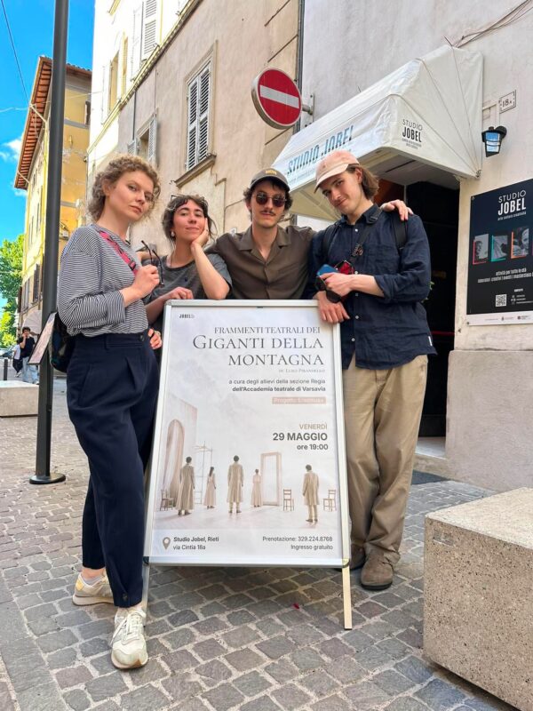 Czworo młodych ludzi pozujących na miejskim deptaku przed wejściem do „Studio Jobel” obok plakatu zapowiadającego spektakl teatralny.